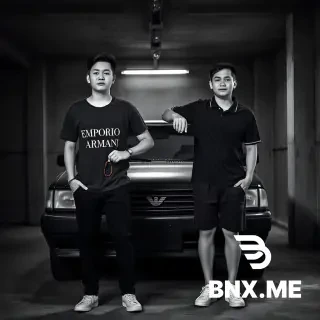 A hyper-realistic, black and white full-body RAW photograph of two 17-year-old teenage boys standing side by side in a dimly lit, gritty garage. In front of them is an old, dusty black cheap sedan car. The lighting is dramatic and moody, coming from a single overhead fluorescent tube, creating hard shadows on the concrete floor.  Left Boy: Tall, lean build, completely clean-shaven (no facial hair, smooth skin). He is wearing a dark grey cotton t-shirt with "EMPORIO ARMANI" written in bold, white, capital letters centered on the chest. He also wears black trousers, a simple bracelet on his wrist, and holds Muslim prayer beads (tesbih) loosely in his right hand. His arms are completely hairless.  Right Boy: Slightly taller than the left boy, also completely clean-shaven (no facial hair at all). He is wearing a black polo shirt (collar popped or flat), black shorts, and clean white sneakers. He has his arm placed casually and protectively around the left boy's shoulder.  Technical Settings: Shot on 35mm film, Canon EOS R5, natural pose, looking towards the camera, serious expressions. 8K resolution, high contrast, sharp focus on the subjects, slightly blurred gritty background. Professional lighting, realistic skin texture and pores. --ar 4:3