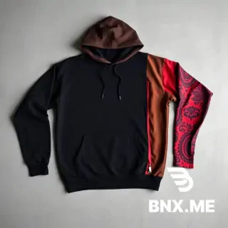 "High-quality studio product shot of a heavyweight, oversized streetwear hoodie laid flat on a neutral grey concrete background. The hoodie features an asymmetrical, vertical color-blocked design: the left side and most of the front are deep matte black, while a contrasting vertical panel on the right side and the right sleeve are a rich chocolate brown. A key detail is a long vertical slit or distressed 'tear' on the outer forearm of the brown sleeve, revealing a secondary inner layer of bright red fabric with an intricate black graphic or paisley-style print. The hoodie has a large front kangaroo pocket, dropped shoulders, and a thick, structured double-layered hood without drawstrings."