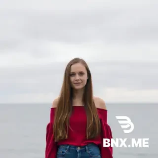 A woman with long, reddish-brown hair, fair skin, standing in front of a body of water (ocean or large lake) under a cloudy sky. She wears an off-the-shoulder red top and blue jeans. The mood is relaxed and outdoorsy.