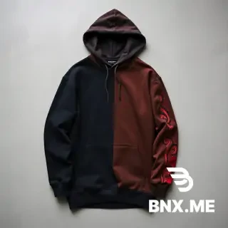 "High-quality studio product shot of a heavyweight, oversized streetwear hoodie laid flat on a neutral grey concrete background. The hoodie features an asymmetrical, vertical color-blocked design: the left side and most of the front are deep matte black, while a contrasting vertical panel on the right side and the right sleeve are a rich chocolate brown. A key detail is a long vertical slit or distressed 'tear' on the outer forearm of the brown sleeve, revealing a secondary inner layer of bright red fabric with an intricate black graphic or paisley-style print. The hoodie has a large front kangaroo pocket, dropped shoulders, and a thick, structured double-layered hood without drawstrings."