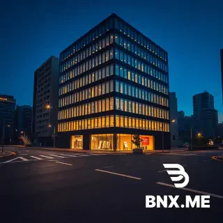 A professional architectural photograph taken at night, capturing a modern, geometric, corner office and residential building. The building features a multi-tiered, stepped facade with sharp, angled corners and a grid of well-lit glass windows, creating a distinct glowing crystal-like effect against the dark blue twilight sky. The ground floor retail space is warmly lit, showcasing visible shelving, a reception counter, and small display cases, possibly a boutique or a curated store. The building is flanked by older, darker, traditional Japanese multi-story buildings and apartment blocks, creating a contrast. In the foreground, a large, empty, dark asphalt road intersection with street markings stretches out. A few empty sidewalks with street lamps and small, manicured trees line the scene. The overall color palette is a deep indigo blue sky and dark asphalt, with warm white and golden light radiating from the modern building's windows. The perspective is a wide-angle shot looking up at the building from the across the street, creating a strong sense of presence and scale. The atmosphere is quiet and still. A tiny, subtle star logo is visible in the extreme bottom-right corner.