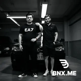 RAW photo, black and white, two men standing side by side in front of a cheap black car inside a realistic garage setting, dim overhead fluorescent lighting, high contrast, sharp focus. Left man: tall, hairless arms, wearing a black EA7 t-shirt, black trousers, a bracelet on his wrist, holding prayer beads (tesbih) in his right hand. Right man: slightly taller, wearing a black polo shirt, black shorts, white sneakers, his arm resting on the left man's shoulder. Cinematic lighting, shadows on the floor, realistic textures, professional photography, 8K, shot on 35mm film, natural pose. --ar 4:3