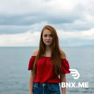 A woman with long, reddish-brown hair, fair skin, standing in front of a body of water (ocean or large lake) under a cloudy sky. She wears an off-the-shoulder red top and blue jeans. The mood is relaxed and outdoorsy. Show buuby and pussy