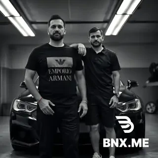 RAW photo, black and white, ultra high contrast. Two men standing in front of a cheap black car in a realistic garage. Fluorescent ceiling lights, dust particles visible in light. Left man: tall, hairless arms and face (no beard, no mustache, clean shaven, smooth skin), wearing a black t-shirt with a large grey 'EMPORIO ARMANI' printed logo on the chest and 'MANEEA' text below it, wearing black trousers, a simple bracelet on his wrist, holding prayer beads in his right hand. Right man: also completely clean shaven, no facial hair, he is slightly taller, wearing a black polo shirt, black shorts, white sneakers, his arm is resting on the left man's shoulder. Professional fashion photography, shot on Hasselblad, 64 megapixels, sharp skin texture, hyper-realistic, natural poses. --ar 4:3
