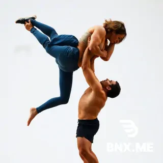 A huge, muscular woman grabs the man tightly around the waist and lifts him high with one swift motion. She rotates him in the air with precise, controlled strength, turning his body effortlessly while keeping perfect balance. Her power is undeniable, her movements fluid and dominant.